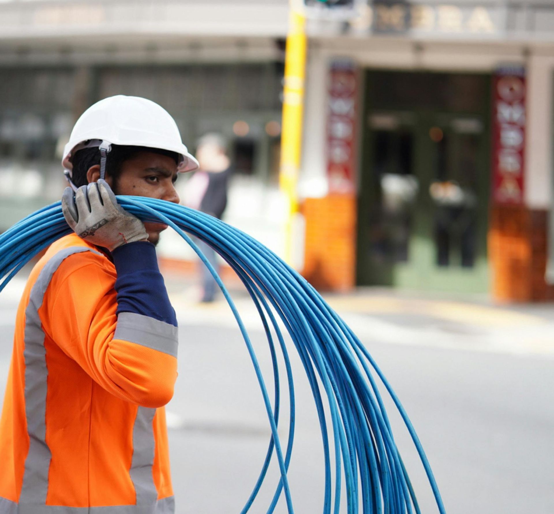 Construction worker in Wellington carrying blue cables, wearing safety gear on a city street.
